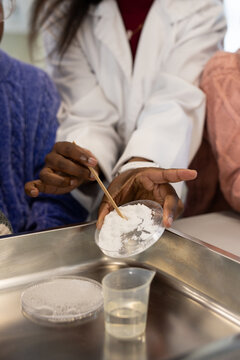 African instructor in lab coat guiding teen girls holding petri dish, stirring powder at lab bench