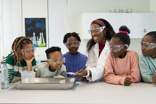 African American educator with schoolkids leading demo at table, in white lab coat, safety goggles