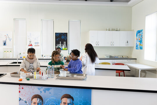 African American teens wearing goggles handling test tubes at bench teacher in lab coat guiding