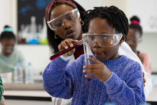 African American teacher guiding child pouring red liquid into tubes at lab wearing coat, goggles