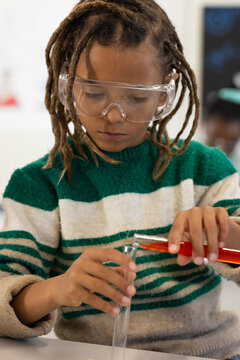 African American child with goggles, striped sweater, pouring orange liquid into test tube in lab