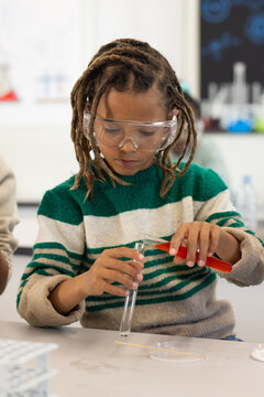 African American preteen pouring red liquid into test tube at lab bench wearing goggles and sweater
