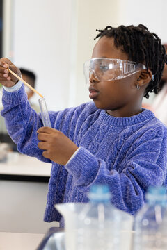 Teen girl wearing goggles and blue sweater, stirring test tube with wooden stick at school lab
