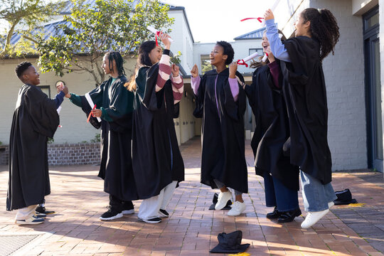 Diverse youth graduates in black gowns jumping and raising red ribboned diplomas in brick courtyard