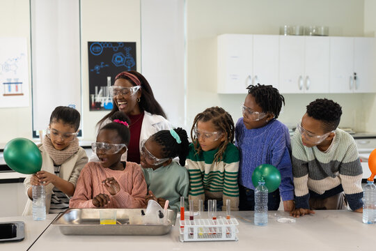 African youth with teacher in lab coat doing experiment at school wearing goggles, using balloons