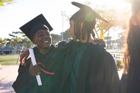 African American teen grads standing on campus in black gowns caps holding red-ribboned diploma