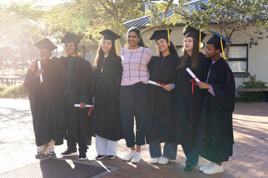 Seven female graduates standing on campus wearing black gowns, holding caps and diplomas, smiling