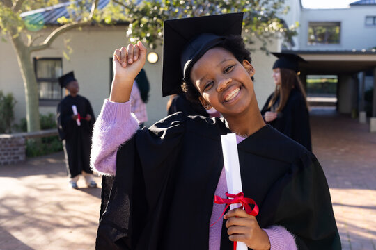 Teenage African American smiling, raising left fist, holding diploma, wearing mortarboard on campus