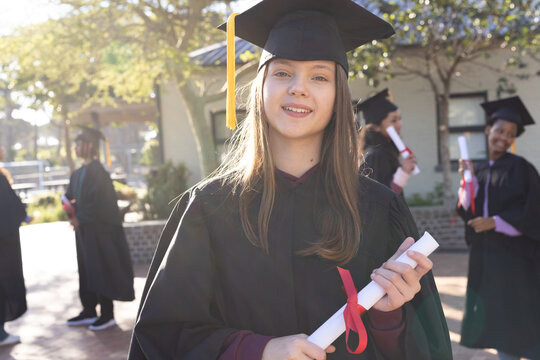 Graduates gathering outdoors, Youth woman center holding red-ribbon diploma in gown and cap