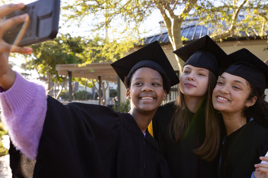 Diverse teen female students taking selfie on campus with smartphone, caps, gowns, tassels