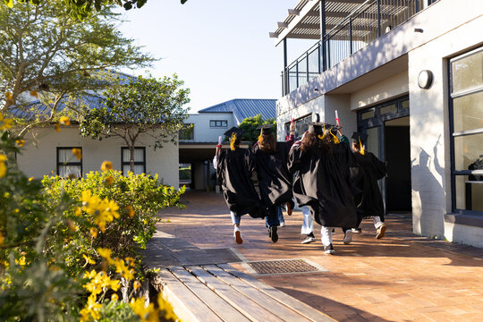 Graduates are running and jumping down brick path on sunlit campus, holding mortarboards, diplomas