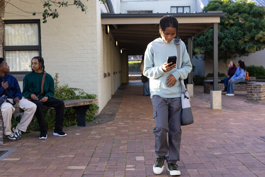 African American teen in hoodie walking down brick courtyard checking phone with tote and earbuds