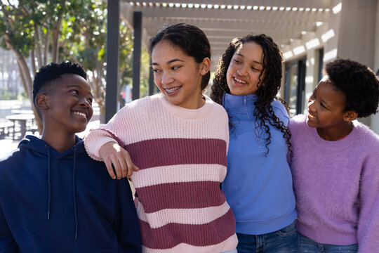 Diverse teenage friends chatting and smiling under pergola on campus with knit sweater, navy hoodie