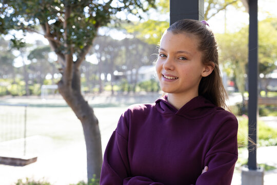 Teenage female standing with arms crossed at park by vertical post and maroon hoodie