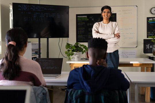 Indian woman instructor presenting to teenage students in classroom, monitor and laptop with code