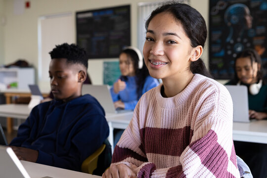 Classmates working at classroom desks, female teenager smiling while using laptop and headphones