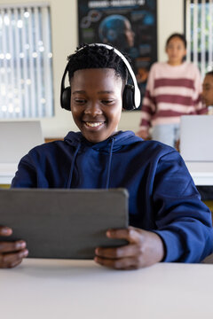 Female teenager smiling, holding tablet, wearing navy hoodie and over-ear headphones at school desk