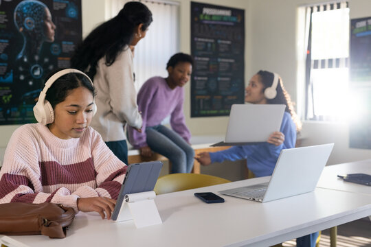 Diverse female students sitting at study table using tablet on white stand wearing headphones