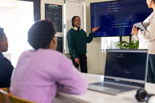 Diverse female students watching African American teen presenting code in classroom with monitor