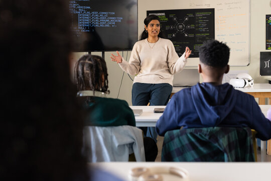 Diverse lab Indian woman teaching code on screen while teens taking notes, VR headset on table