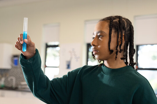 African American teen male wearing green sweatshirt holding test tube examining blue liquid at sink