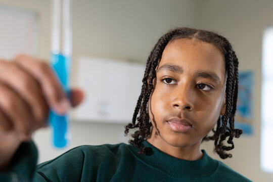African American teen wearing green top holding blue test-tube studying by lab window blurred-hand