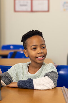 School-age male student sitting at classroom desk wearing knit sweater with blue chairs, posters