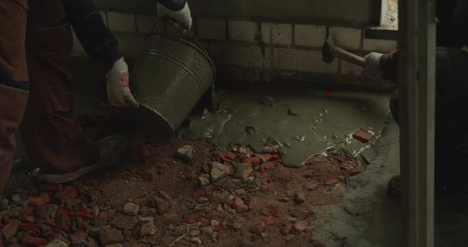 Male builder in uniform pouring liquid cement mortar from a bucket onto a dirty floor with broken bricks during repair work on a construction site, creating a new foundation for the room