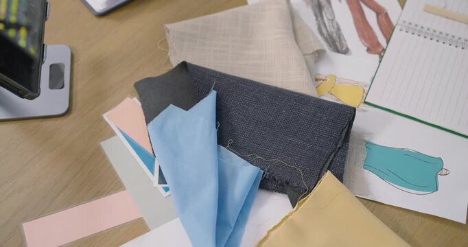 Camera is panning up and left, showing fabric swatches, fashion sketches, notebook for design