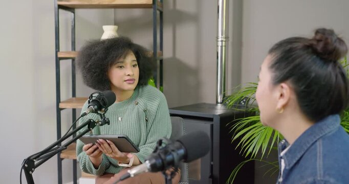 African American host reading tablet and speaking into mic while Asian guest responding, podcast