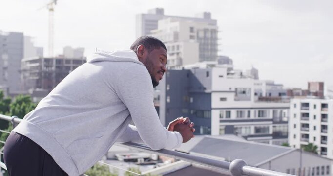 African man in gray hoodie leaning on roof railing shifting, glancing at crane noting construction