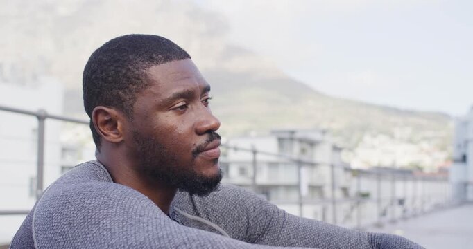 African man sitting roof wearing grey sweater, opening eyes with daylight showing hill over railing