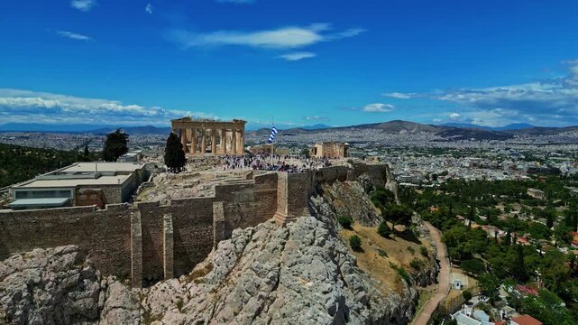 Aerial view of the Acropolis of Athens featuring the ancient Parthenon temple, historic stone walls, and the sprawling city landscape under a bright blue sky in Greece.