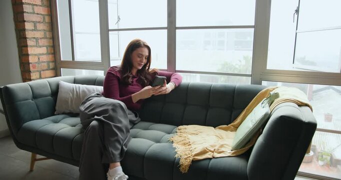 Woman looking at phone, scrolling, leaning back, gazing out window, reading on sofa burgundy-top