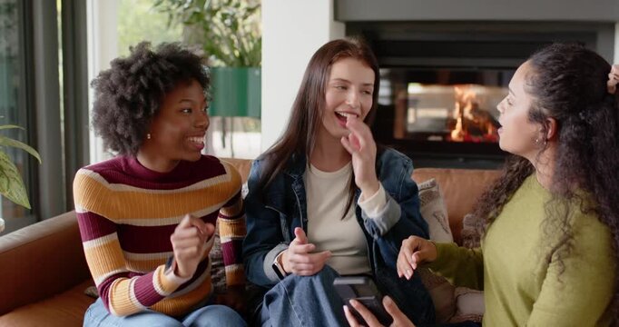 Female friends sitting on couch, rightmost woman holding smartphone sharing clip, others laughing