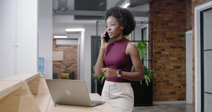 African American woman in maroon top ending smartphone call, typing on laptop to work at reception