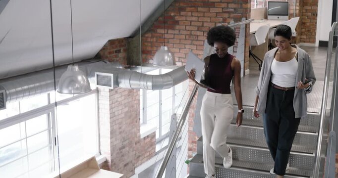 Diverse women coworkers descending stairs loft with laptop and smartphone, screen sparking review