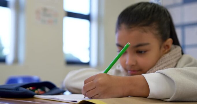 African American girl seeing yellow book, writing with green pencil at school desk, doing classwork