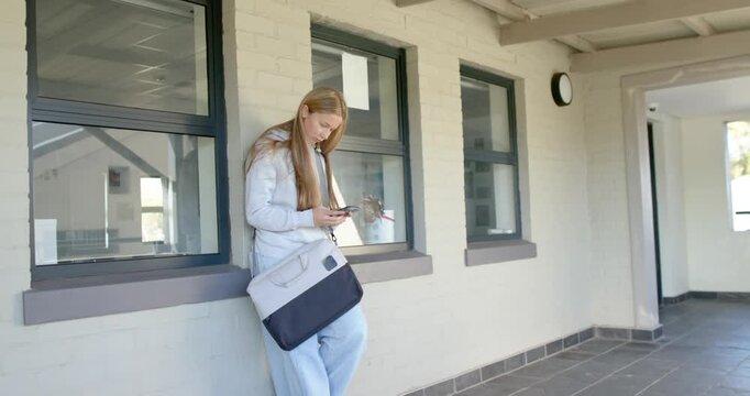 Female youth in puffer jacket leaning on painted wall under porch reading text adjusting tote bag