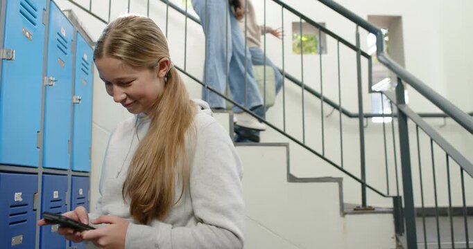 Teenage girl standing at blue lockers on school stairs checking phone reading texts, peers passing