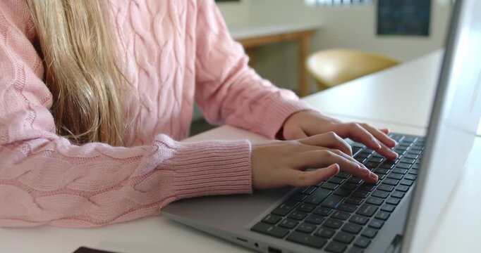 Teenage female in pink knit settling hands on silver laptop at desk, starting typing for task