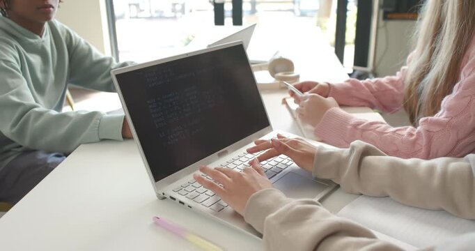 Diverse teenage students typing on white laptop near windows, phone scrolling, working for deadline