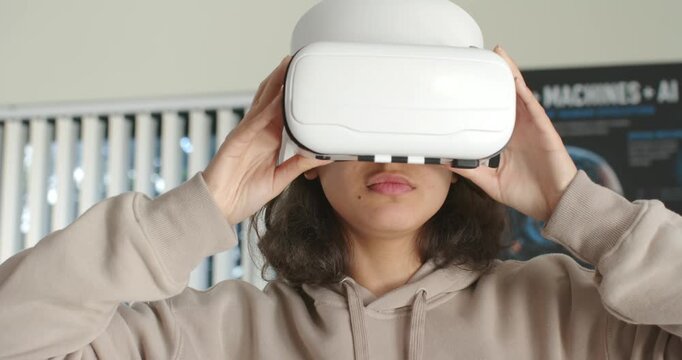African American teenage female in beige hoodie grabbing white VR headset, testing VR in lab