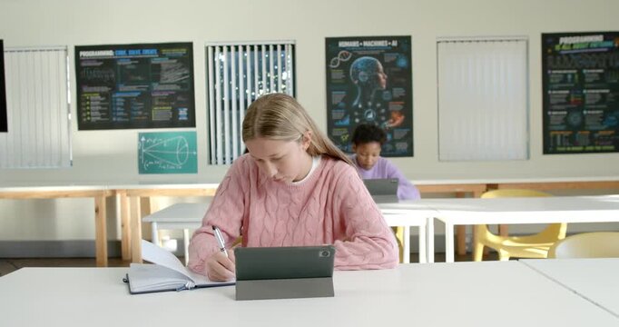 Diverse female students studying in classroom, front teen following tablet and taking notes