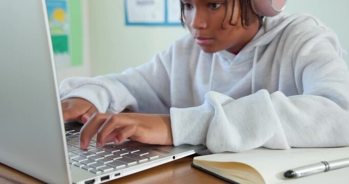 African American teen typing on laptop at desk, wearing pink headphones, using notebook pen