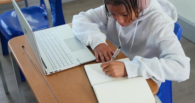 African American teen writing, glancing at laptop, and typing, studying at school with pink headset