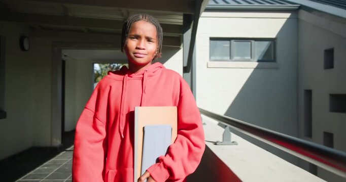 Male youth red hoodie walking from corridor toward railing, squinting in sunlight, holding folders