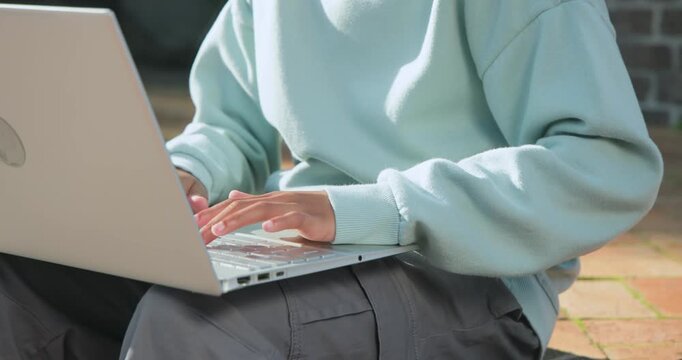 African American teen male starting assignment, typing on laptop, pausing to think on outdoor bench