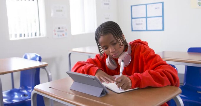 African teen student writing notes at class desk, tapping tablet, raising hand, answering question