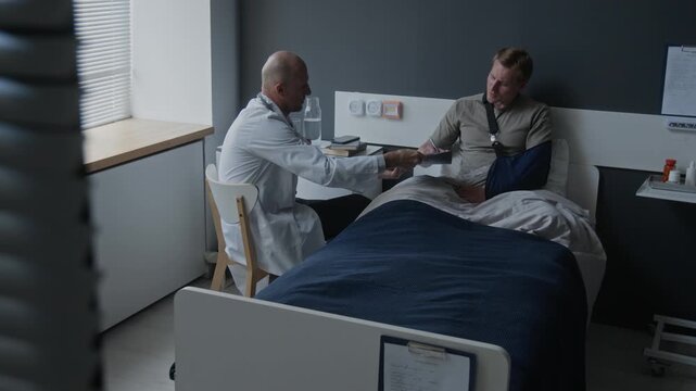 Caucasian male patient with arm sling signing medical document held by doctor in hospital room. Ideal for healthcare insurance, patient consent, and medical services marketing